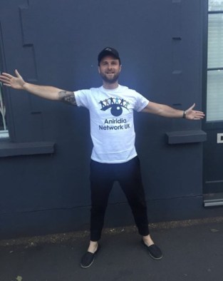 Matt Hill smiling, with arms outstretched, wearing a white Aniridia Network t-shirt, black trousers and a black cap.